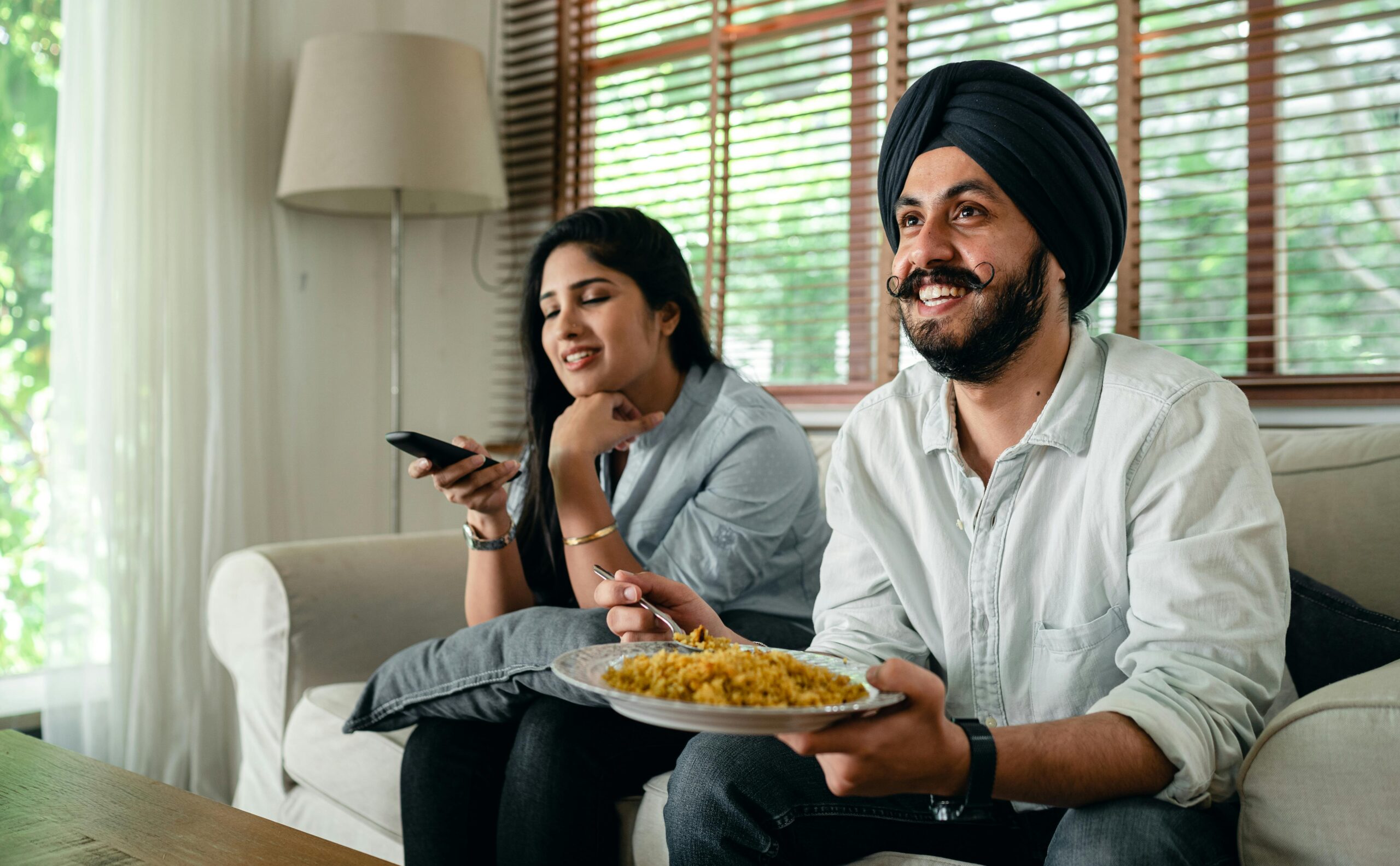 Positive young bearded man in casual wear and turban eating traditional saffron rice on sofa while wife switching channels on TV with remote controller vastu